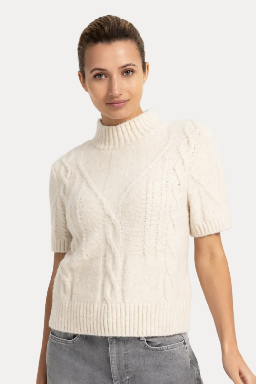 A woman modeling a short-sleeve cable-knit mock neck sweater in cream, paired with gray jeans, against a plain white studio background.