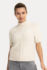 A woman modeling a short-sleeve cable-knit mock neck sweater in cream, paired with gray jeans, against a plain white studio background.