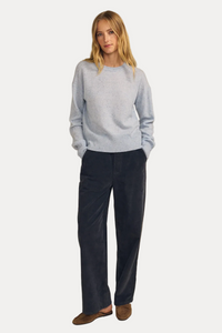 A woman stands against a plain beige studio background wearing a light blue knit sweater, dark navy wide-leg pants, and brown slip-on shoes.