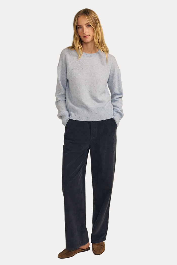 A woman stands against a plain beige studio background wearing a light blue knit sweater, dark navy wide-leg pants, and brown slip-on shoes.