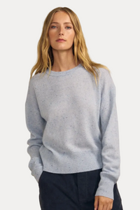 A woman modeling a light blue speckled knit crewneck sweater with dark navy pants, set against a solid beige studio background.