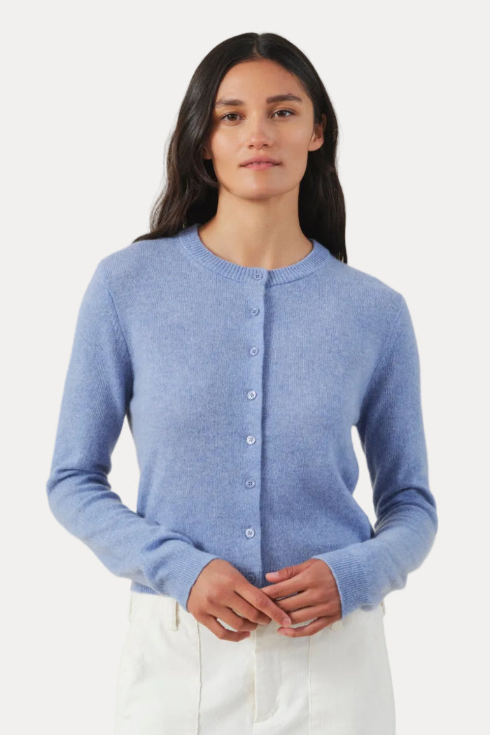 A woman modeling a light blue knit cardigan sweater with white pants, standing against a plain beige studio background.