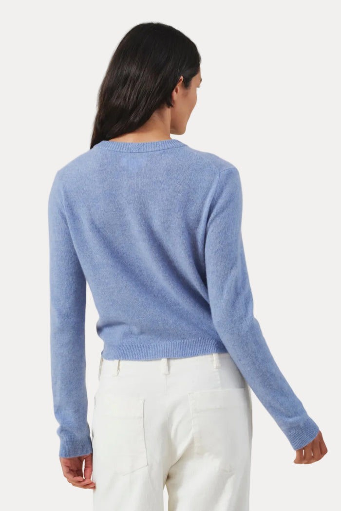 A woman shown from the back wearing a light blue long-sleeve knit sweater and white trousers, standing against a plain light beige studio background.