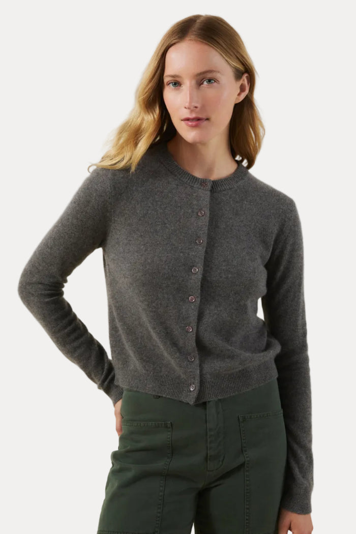 A woman modeling a dark gray button-up cardigan sweater paired with dark green pants, set against a plain beige studio background.