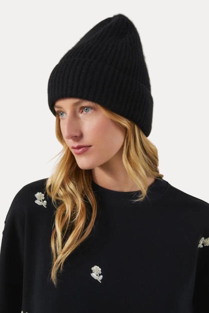 A woman wearing a black ribbed knit beanie and a black crewneck sweatshirt with small white embroidered floral details, photographed against a plain light beige studio background.