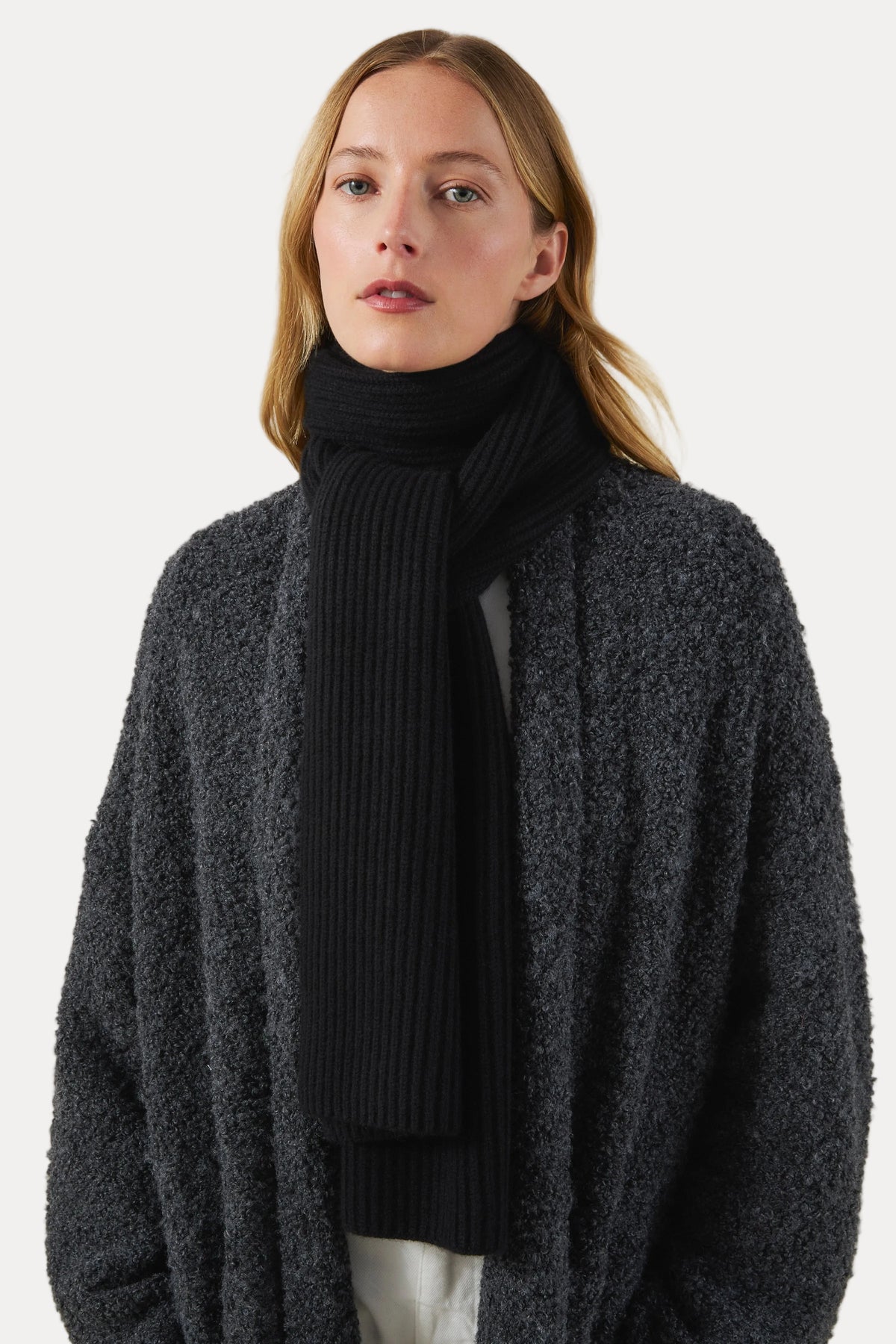 A woman wearing a dark charcoal gray bouclé cardigan and a black ribbed turtleneck scarf, photographed against a plain light beige studio background.