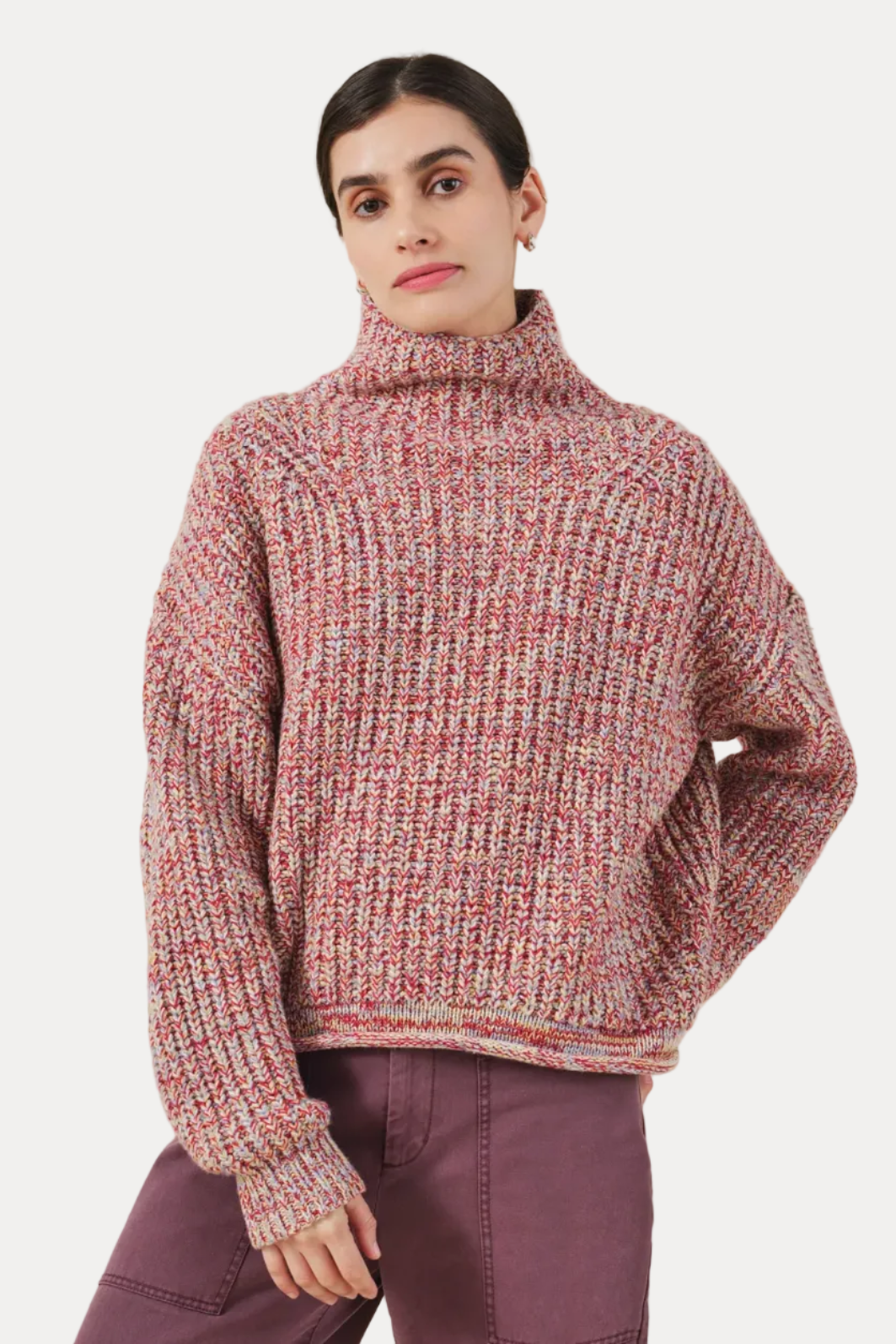 A woman wearing a chunky knit turtleneck sweater in a heathered pink, red, and cream marled yarn, paired with mauve pants, against a solid light beige background.