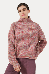 A woman wearing a chunky knit turtleneck sweater in a heathered pink, red, and cream marled yarn, paired with mauve pants, against a solid light beige background.