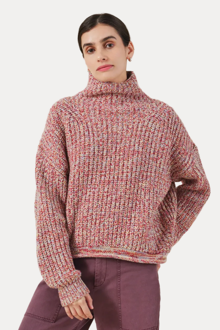 A woman wearing a chunky knit turtleneck sweater in a heathered pink, red, and cream marled yarn, paired with mauve pants, against a solid light beige background.