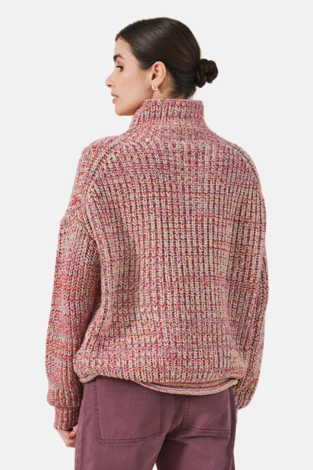 A woman shown from the back wearing a chunky knit mock-neck sweater in a heathered pink, red, and beige marled pattern, paired with mauve pants, against a solid light beige background.