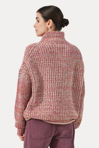 A woman shown from the back wearing a chunky knit mock-neck sweater in a heathered pink, red, and beige marled pattern, paired with mauve pants, against a solid light beige background.