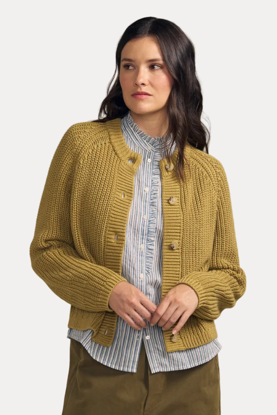 A woman modeling a mustard yellow ribbed knit cardigan with brown buttons, worn over a blue and white striped shirt and paired with olive green pants, against a solid beige background.