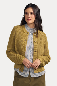 A woman modeling a mustard yellow ribbed knit cardigan with brown buttons, worn over a blue and white striped shirt and paired with olive green pants, against a solid beige background.
