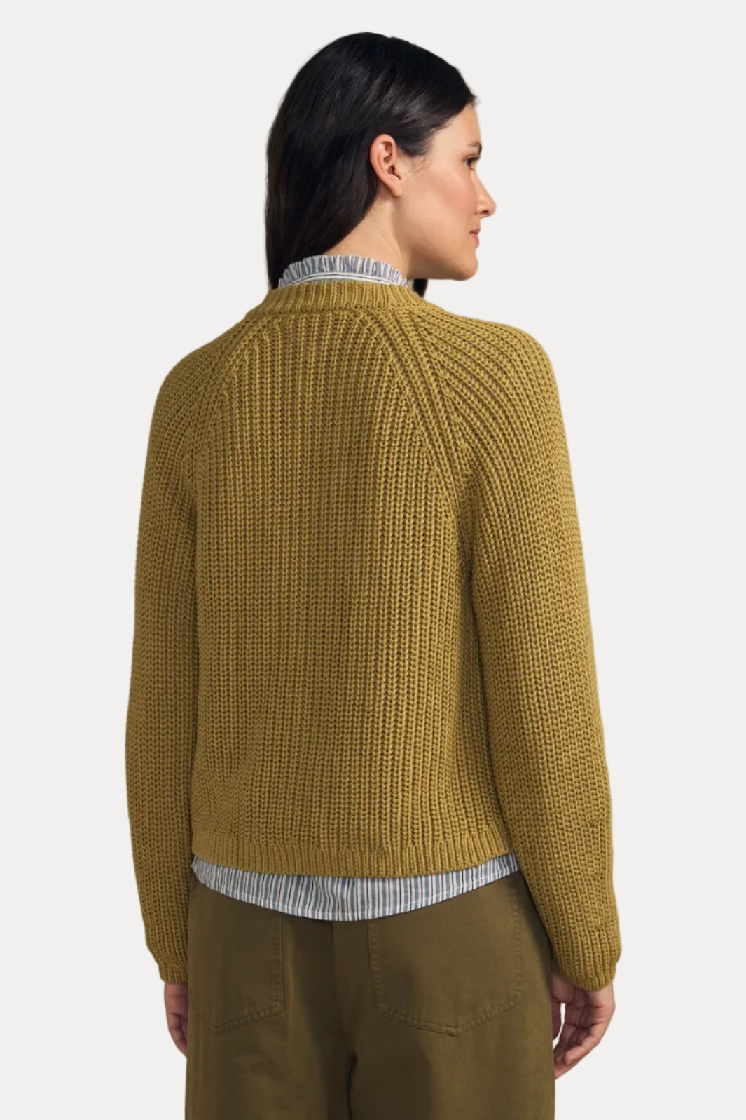 A woman shown from the back wearing a textured mustard yellow knit sweater over a striped shirt and olive pants, set against a solid beige background.