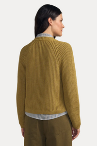 A woman shown from the back wearing a textured mustard yellow knit sweater over a striped shirt and olive pants, set against a solid beige background.