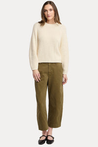 A woman modeling a cream ribbed knit sweater and olive green cropped trousers against a light gray studio background.