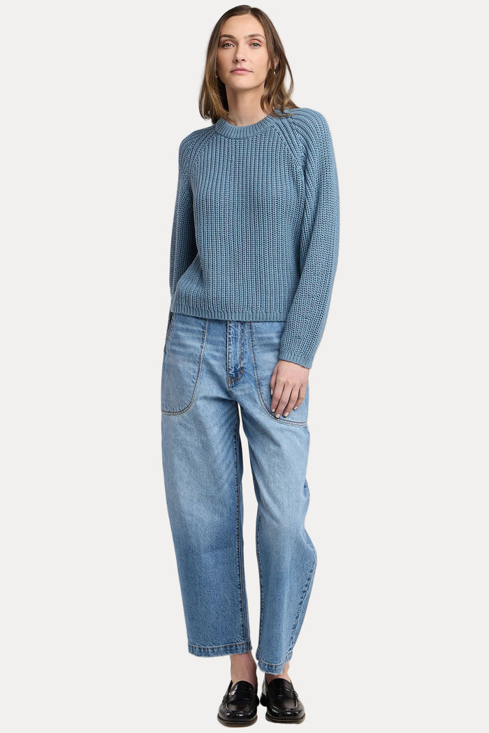 A woman modeling a blue ribbed knit sweater and light blue wide-leg jeans against a plain light gray studio background.