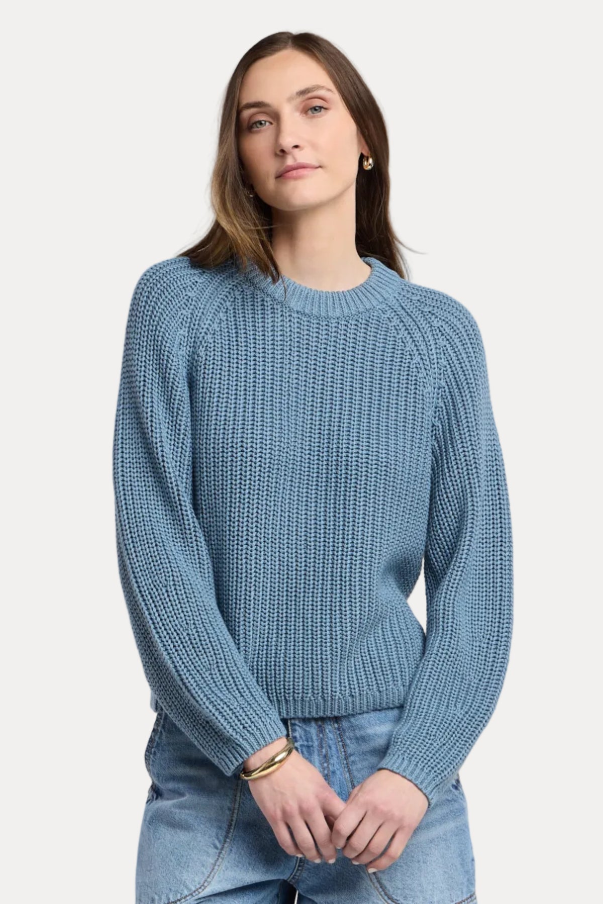 A woman wearing a textured blue knit crewneck sweater and light wash blue jeans, standing against a neutral light beige studio background.