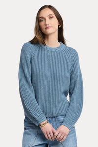 A woman wearing a textured blue knit crewneck sweater and light wash blue jeans, standing against a neutral light beige studio background.