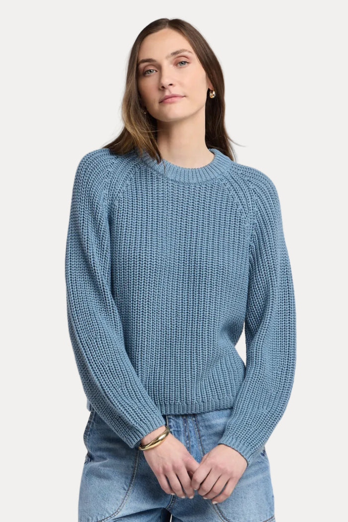 A woman wearing a textured blue knit crewneck sweater and light wash blue jeans, standing against a neutral light beige studio background.