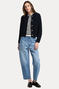 A woman modeling a dark navy ribbed knit cardigan with gold buttons over a black and white striped top, paired with light blue cropped jeans and black loafers, against a light beige studio background.