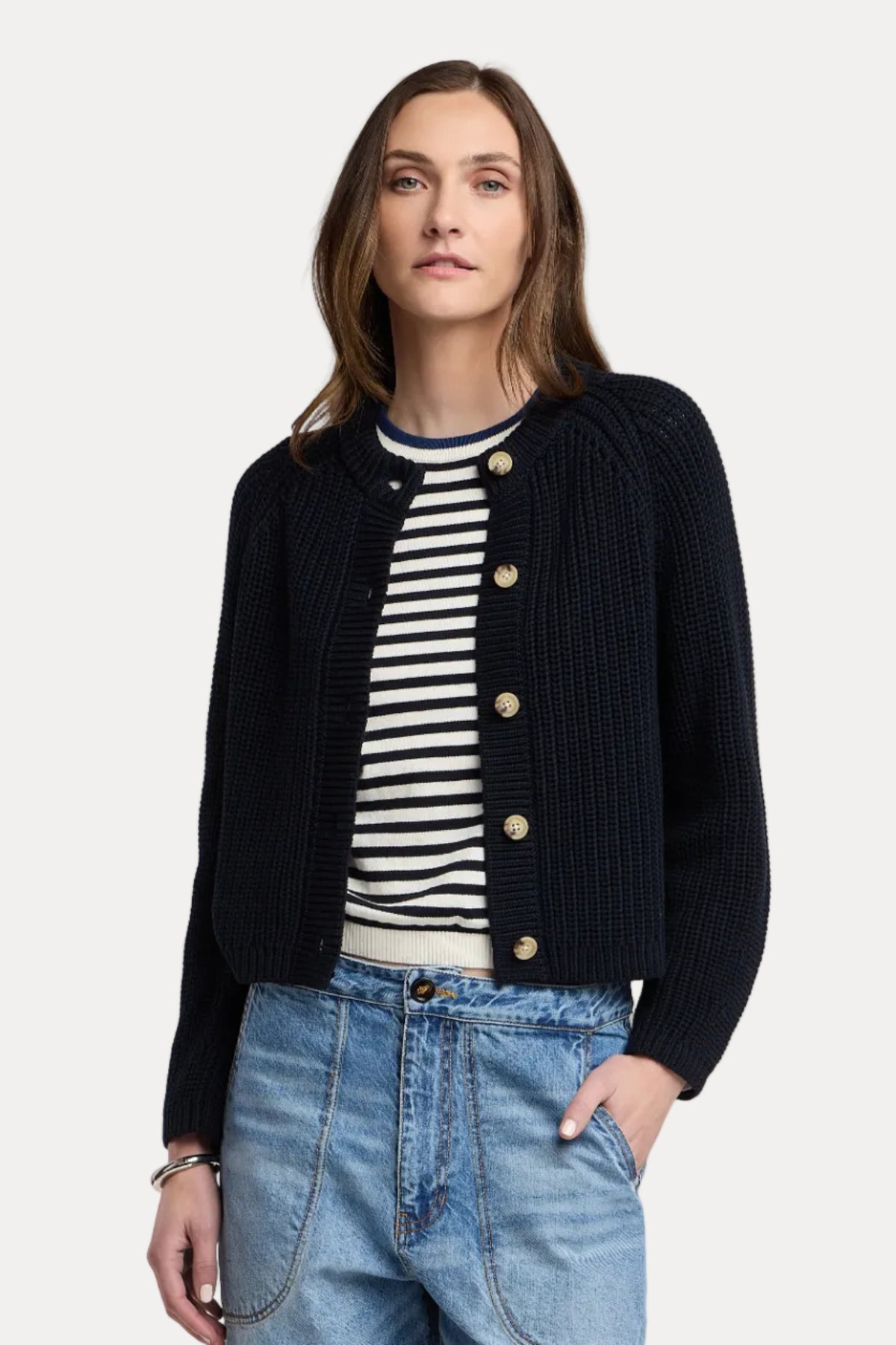 A woman modeling a dark navy blue ribbed knit cardigan with gold buttons, worn over a black and white striped shirt and paired with light blue jeans, against a solid light beige background.