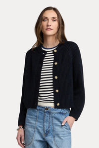 A woman modeling a dark navy blue ribbed knit cardigan with gold buttons, worn over a black and white striped shirt and paired with light blue jeans, against a solid light beige background.