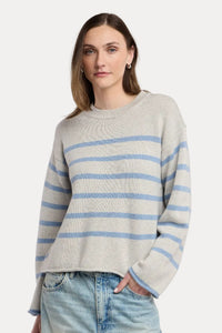A woman wearing a light gray knit sweater with horizontal light blue stripes and light-wash blue jeans, standing against a plain light beige studio background.