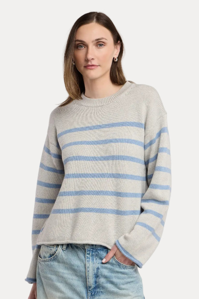 A woman wearing a light gray knit sweater with horizontal light blue stripes and light-wash blue jeans, standing against a plain light beige studio background.