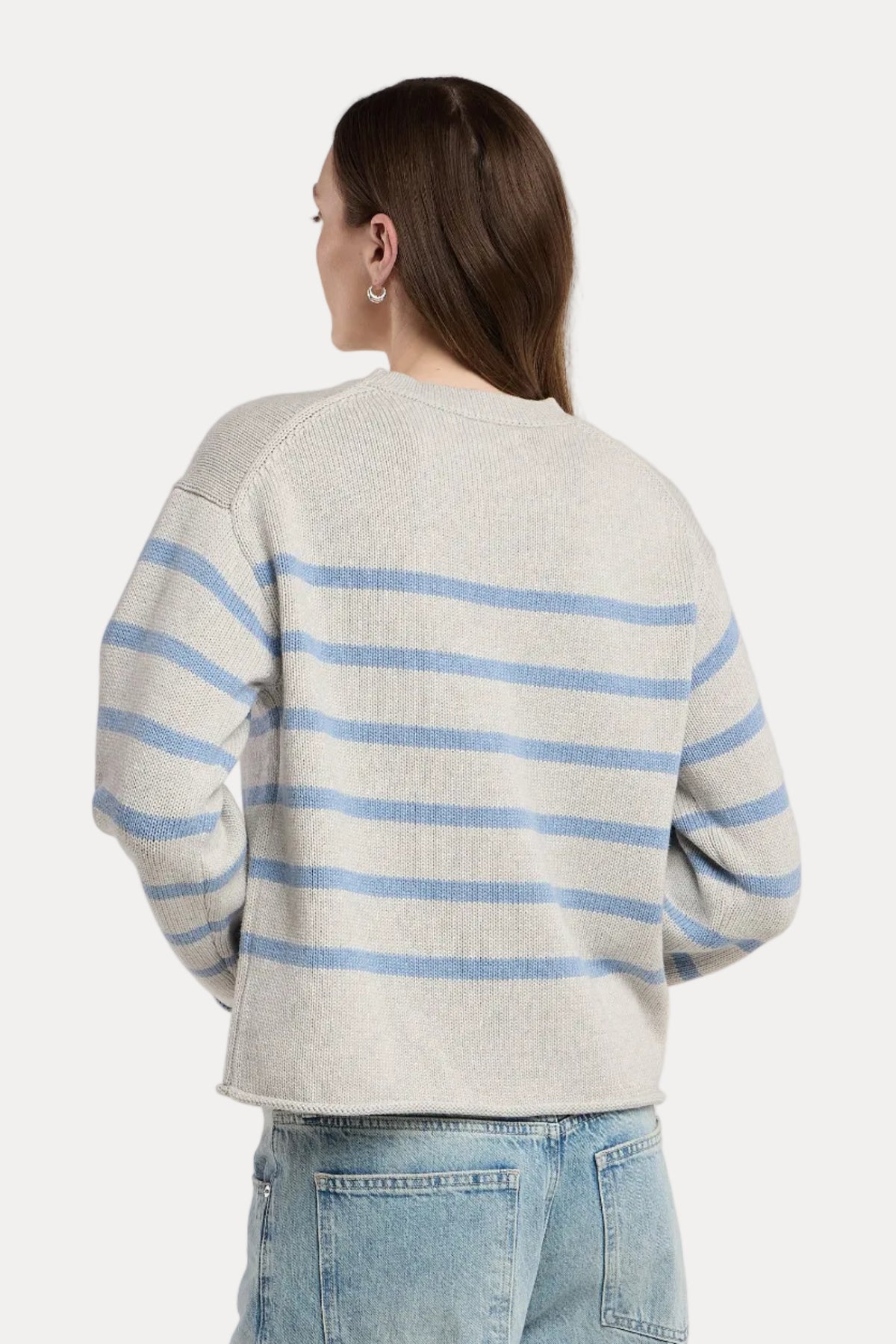 A woman shown from the back wearing a light gray knit sweater with horizontal light blue stripes and light-wash blue jeans, photographed against a plain light gray studio background.