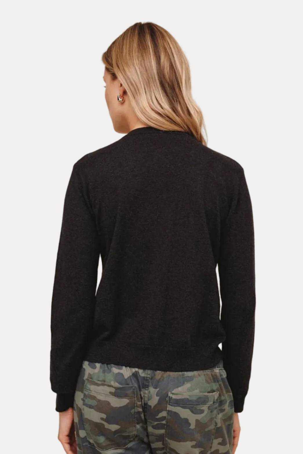 A woman shown from the back wearing a dark charcoal heather knit sweater and camouflage pants, standing against a solid beige studio background.