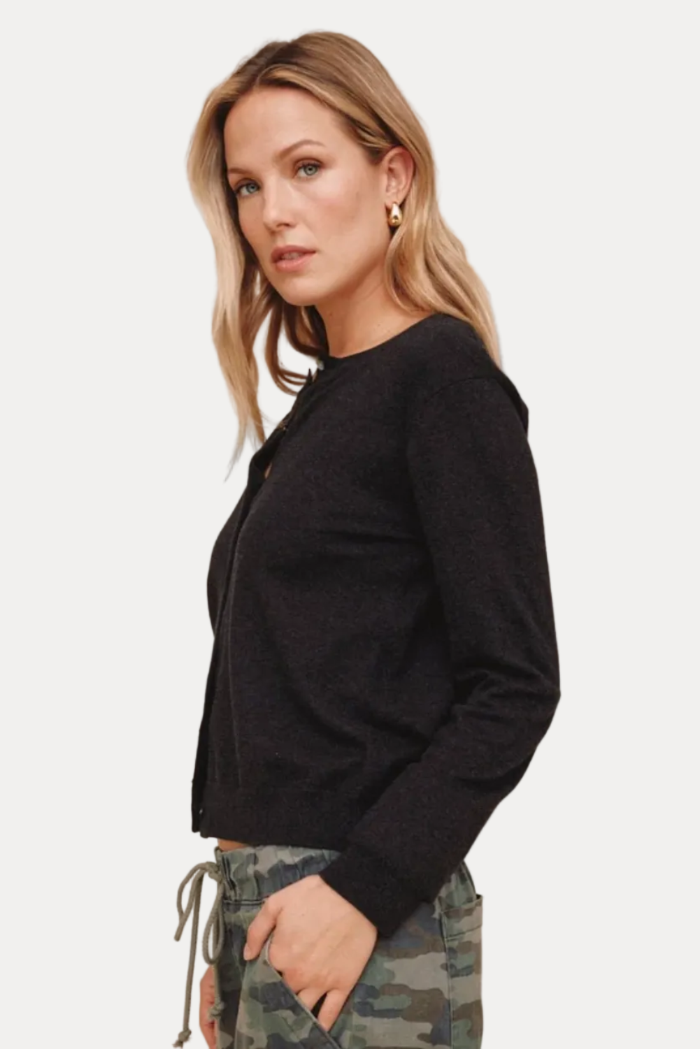 A woman wearing a black long-sleeve knit cardigan with a buttoned front, paired with camouflage pants, photographed against a solid beige studio background.