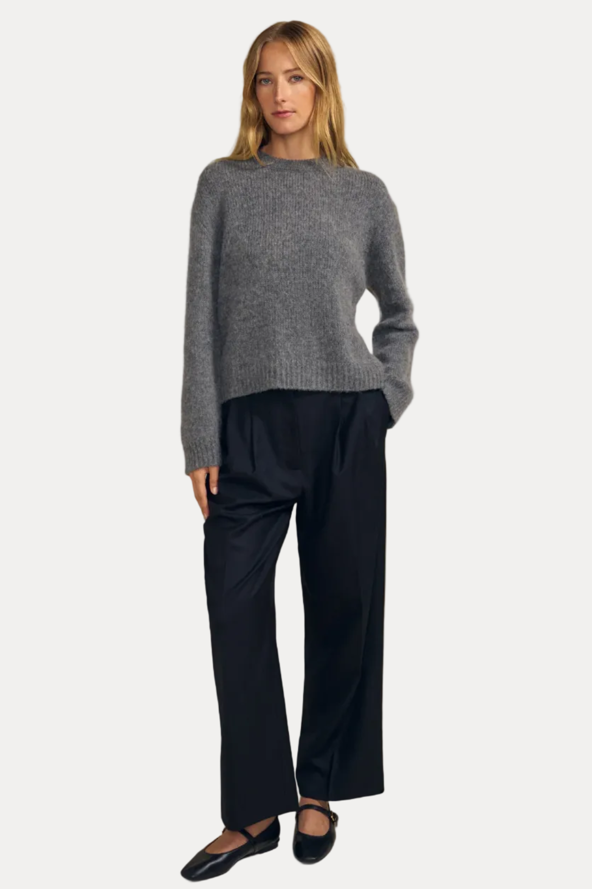 A woman modeling a gray knit sweater and black wide-leg trousers against a solid beige studio background.
