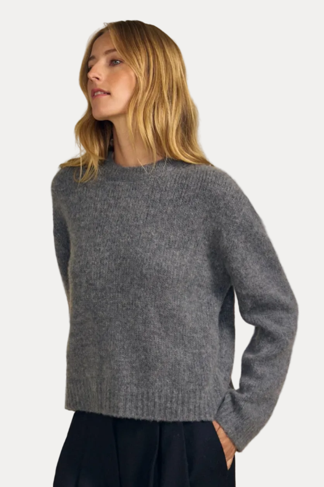 A woman wearing a heather gray knit crewneck sweater and dark navy trousers, standing against a solid beige studio background.