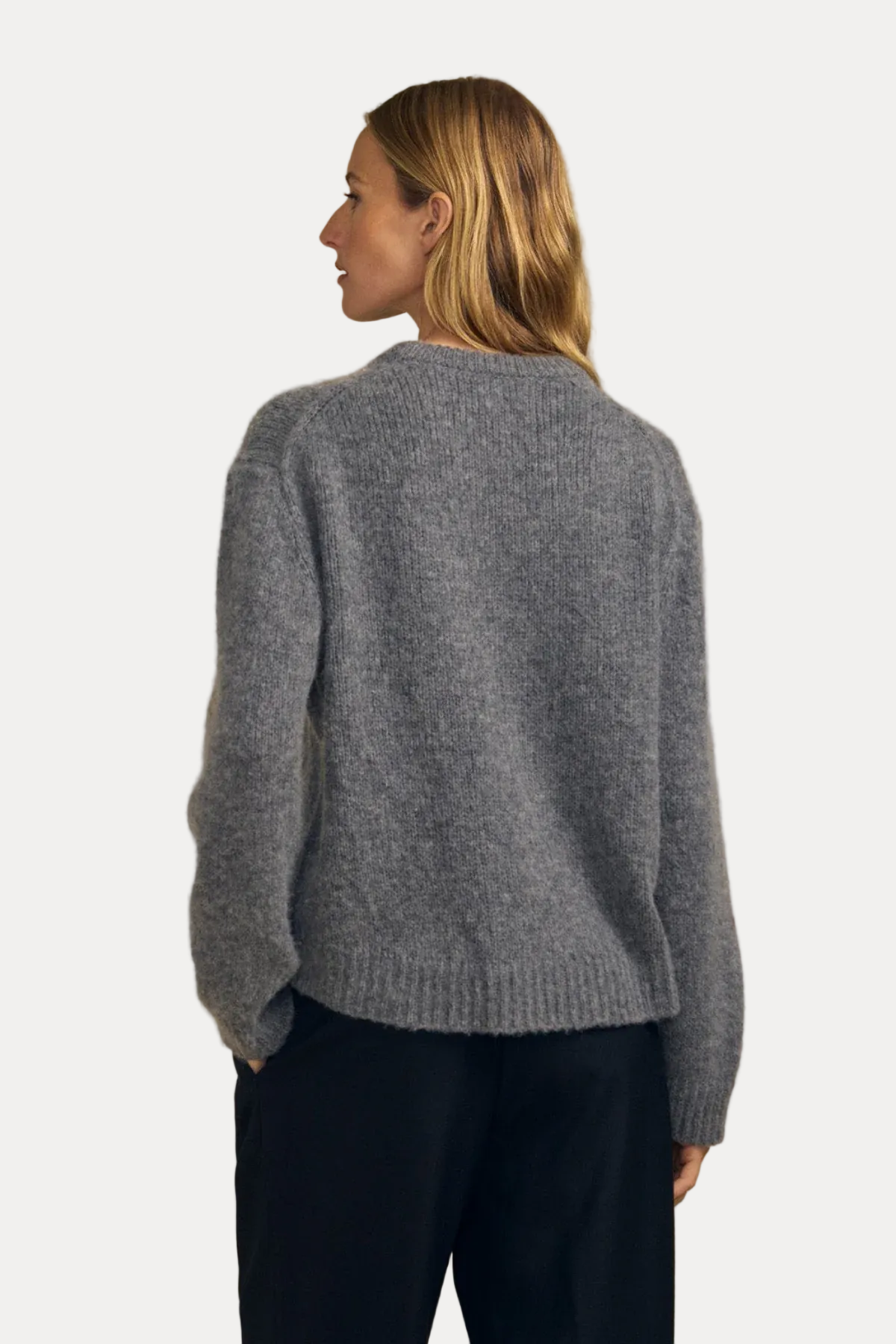 A woman shown from the back wearing a heather gray knit sweater and black pants, standing against a plain beige studio background.