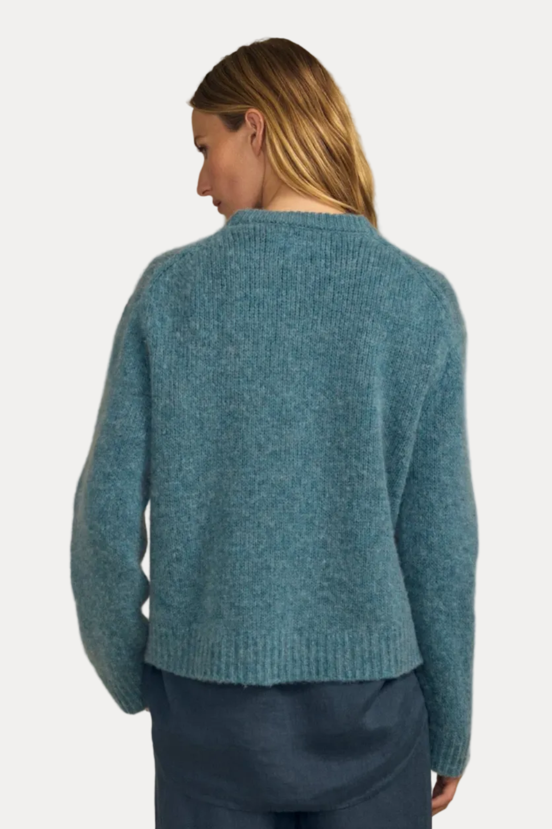 A woman shown from the back wearing a fuzzy knit sweater in a muted teal-blue color, paired with dark blue pants, against a solid beige background.