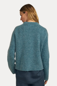 A woman shown from the back wearing a fuzzy knit sweater in a muted teal-blue color, paired with dark blue pants, against a solid beige background.