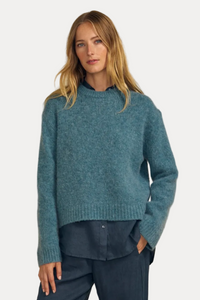 A woman wearing a textured, heathered teal blue knit sweater with a crew neck, paired with dark blue pants, set against a solid beige background.