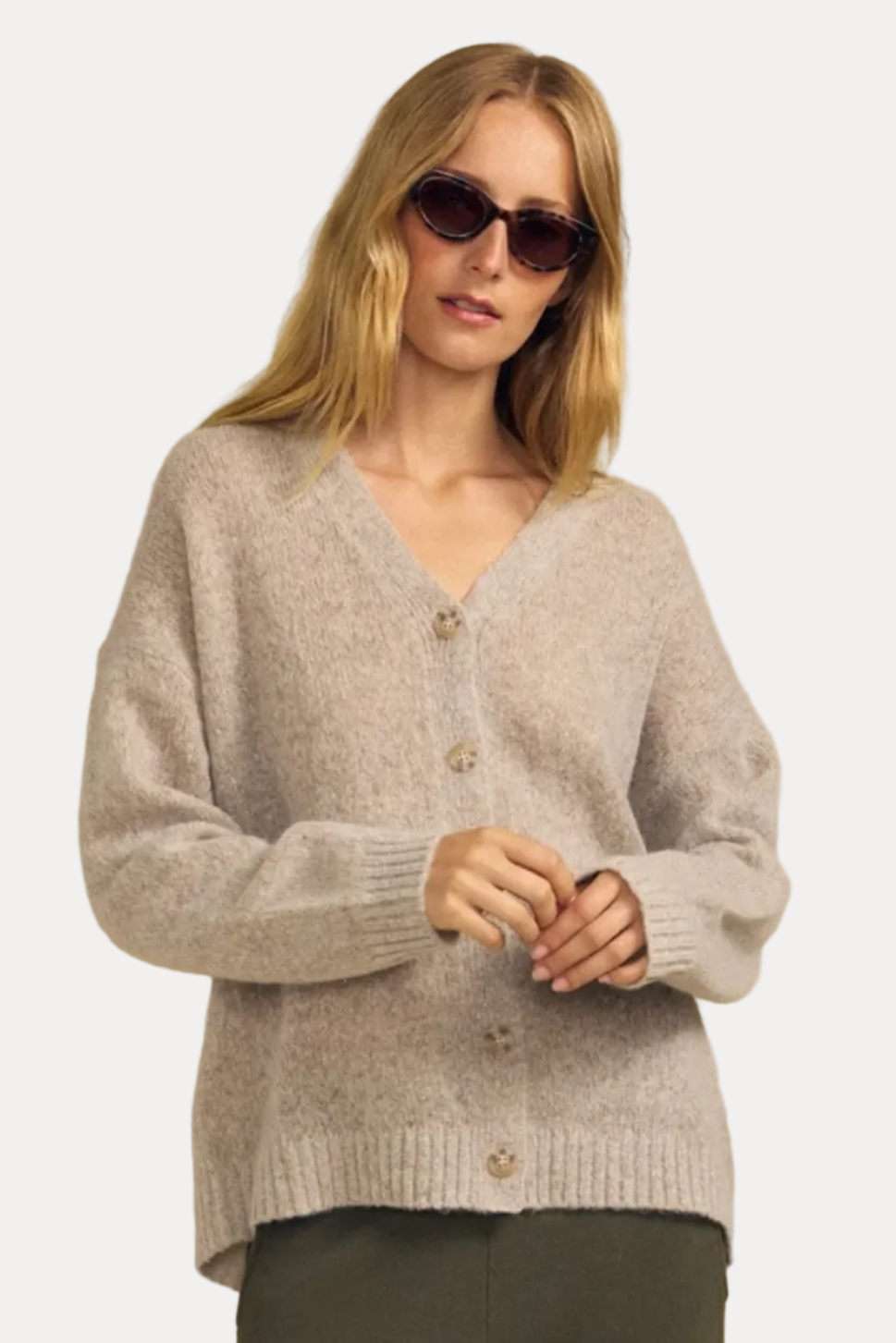 A woman modeling a light beige heathered knit cardigan sweater with brown buttons, paired with dark olive pants, against a solid beige background.