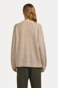 A woman viewed from behind wearing a light beige textured knit sweater and dark olive green pants, set against a solid warm beige background.