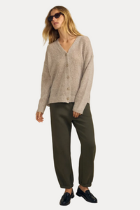 A woman modeling a light beige knit cardigan sweater, olive green trousers, and black loafers against a solid beige studio background.