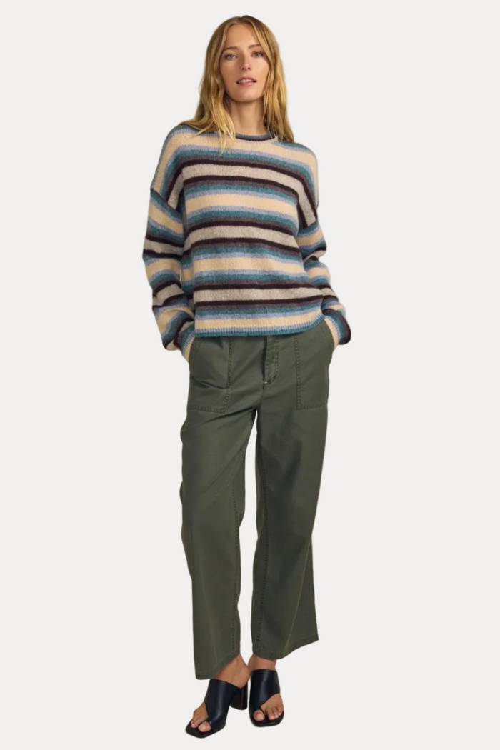 A woman stands against a plain beige studio background wearing a fuzzy striped knit sweater with blue, beige, and brown tones, olive green cropped pants, and dark heeled sandals.