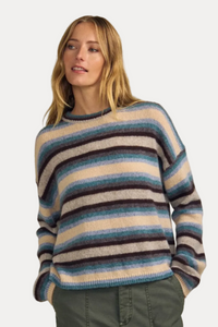 A woman wearing a fuzzy striped knit sweater with horizontal bands of blue, beige, and dark brown, paired with olive green pants, set against a solid beige background.
