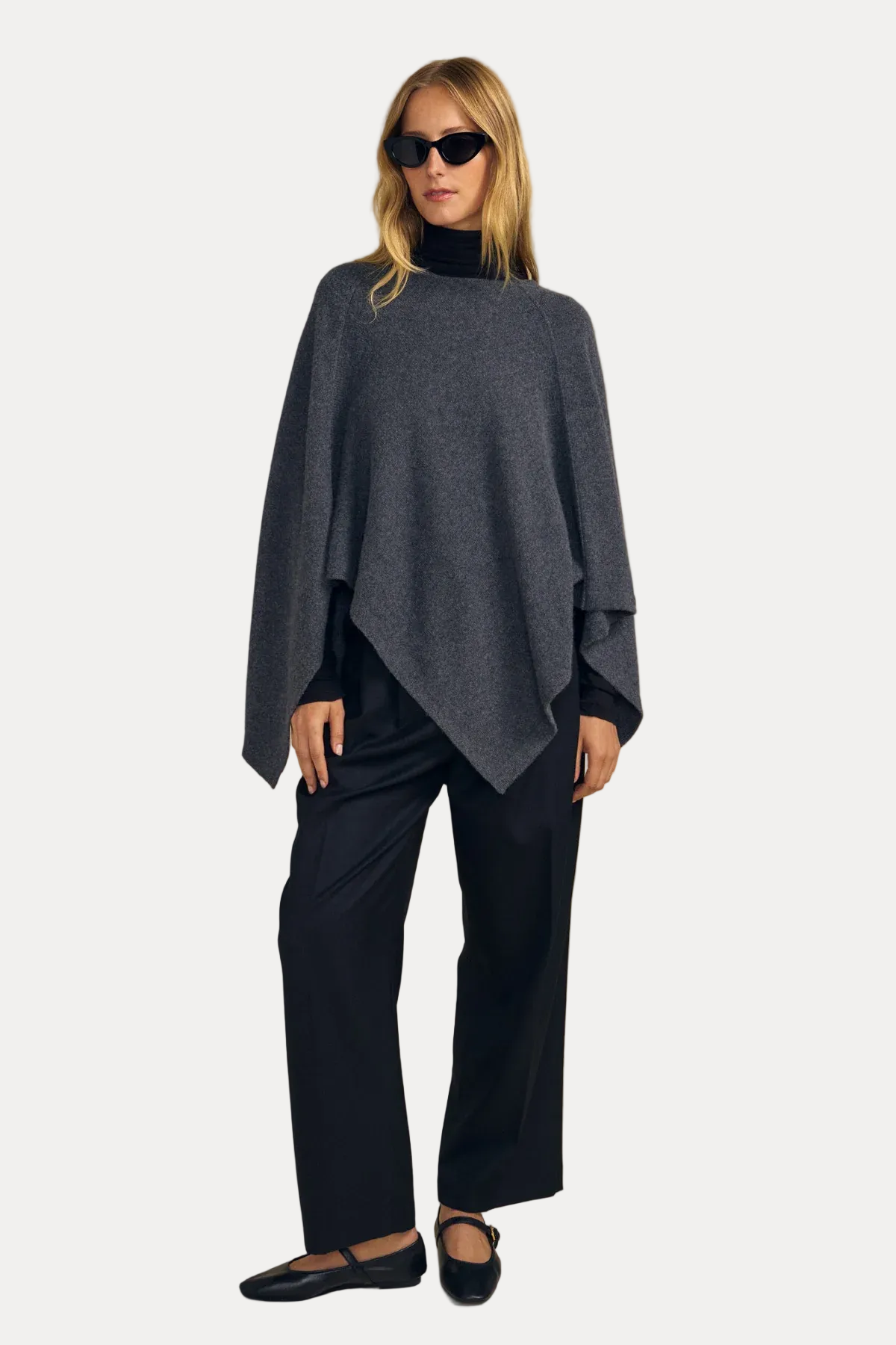 A woman modeling a dark gray draped poncho sweater over a black turtleneck, paired with black wide-leg trousers and black flats, against a solid beige studio background.