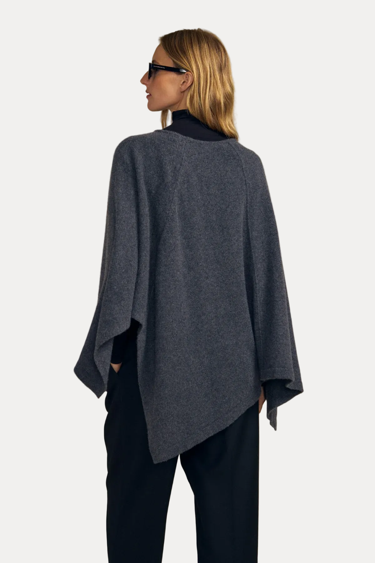 A woman shown from the back wearing a dark gray heathered poncho-style top over a black turtleneck and black pants, standing against a solid beige studio background.