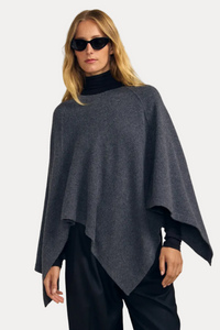 A woman modeling a dark gray knit poncho over a black turtleneck and black wide-leg pants, standing against a solid beige studio background.