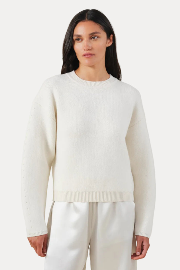 A woman modeling a cream-colored ribbed knit crewneck sweater and matching cream wide-leg trousers against a solid light beige background.