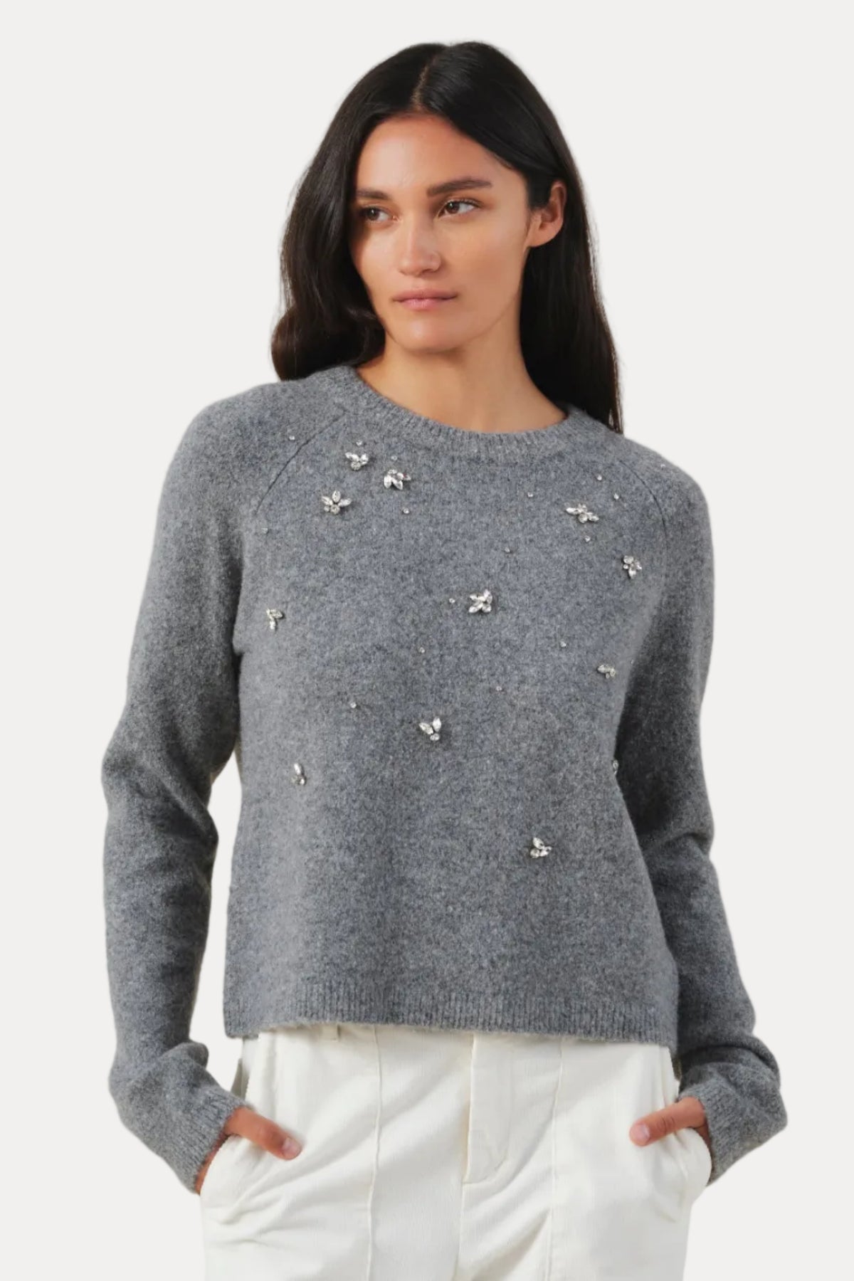 A woman modeling a gray knit sweater with small silver floral appliqués, paired with white pants, against a plain beige studio background.