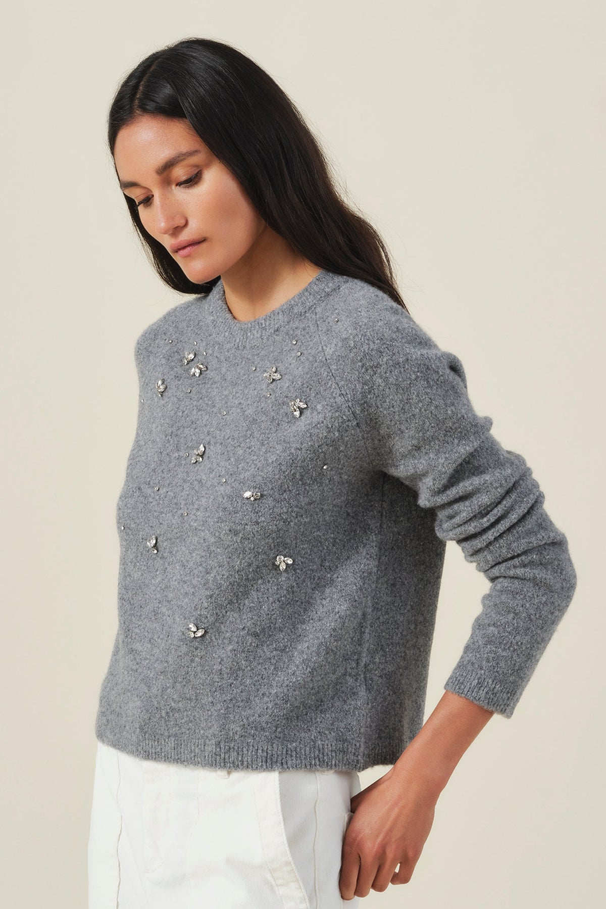 A woman wearing a gray knit sweater with small metallic floral embellishments and white trousers, photographed against a plain light beige studio background.