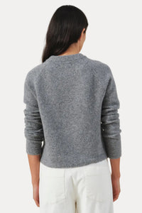 A woman viewed from the back wearing a heather gray long-sleeve knit sweater and white pants, standing against a plain light beige studio background.